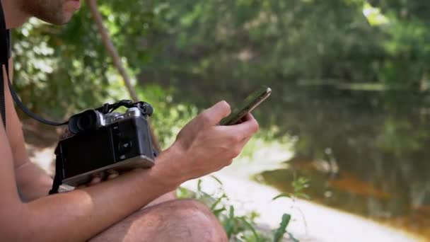 Ένας φωτογράφος κρατάει κάμερα, Smartphone, κάθεται σε μια όχθη του ποταμού. 4K — Αρχείο Βίντεο