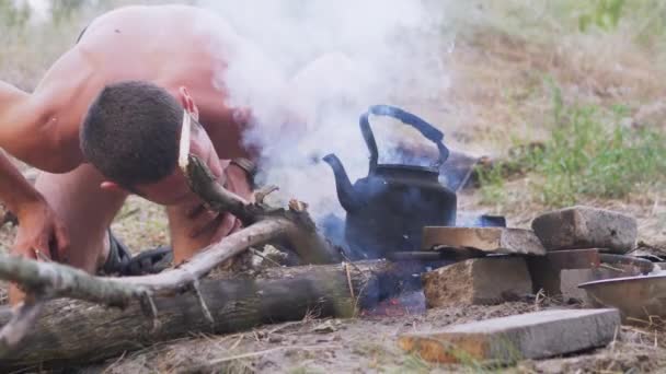 Αρσενικό είναι Fanning the Bonfire Outdoors με ένα βραστό βραστήρα πάνω από τη φωτιά. 4K. — Αρχείο Βίντεο