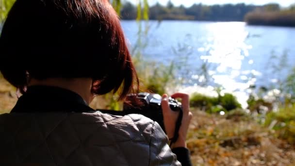 Flicka fotograferar ett höstlandskap, står vid vattnet i solens strålar. 4K — Stockvideo