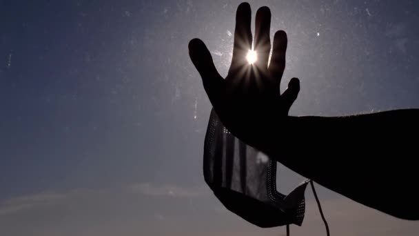 Mão feminina com uma máscara na palma da mão tocando uma janela suja, estende-se para raios de sol — Vídeo de Stock