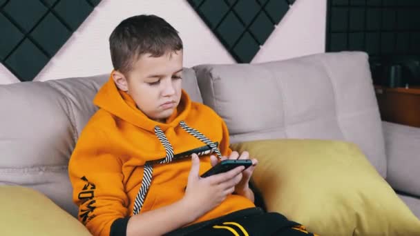 Nastolatek w żółtym kapturze siedzi na kanapie Gra mobilna na smartfonie w domu — Wideo stockowe