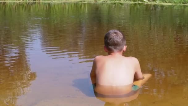 Bakifrån av ett barn som sitter på sand upp till midjan i en genomskinlig, grund flod — Stockvideo