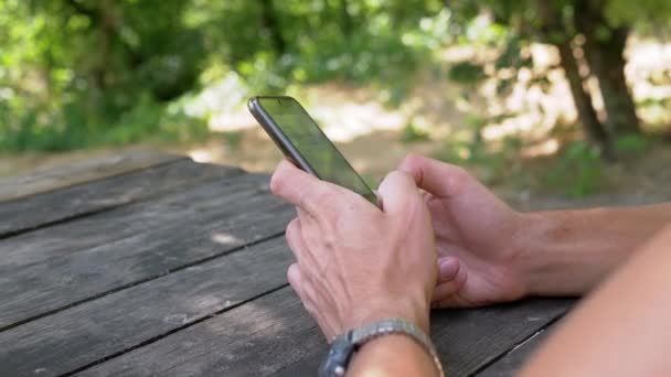 Um macho segura um smartphone nas mãos, sentado em uma mesa de madeira de verão na natureza — Vídeo de Stock