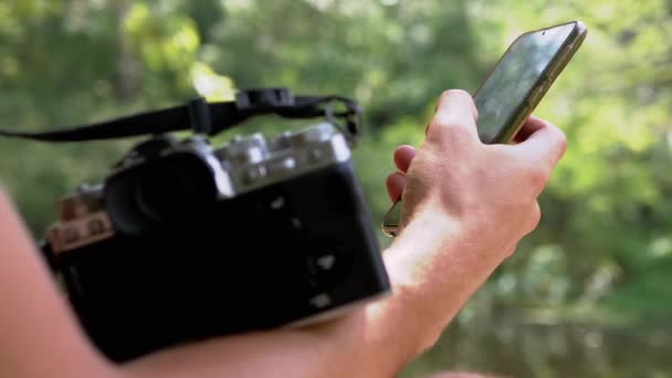 Ένας φωτογράφος κρατάει κάμερα, Smartphone, κάθεται σε μια όχθη του ποταμού. Εστίαση — Αρχείο Βίντεο