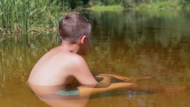 Uma criança senta-se na areia até a cintura em um rio transparente e raso. 4K — Vídeo de Stock