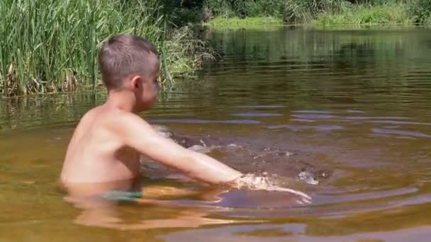 Un niño triste salpica en un río transparente y poco profundo al atardecer. 4K. De cerca. — Vídeo de stock