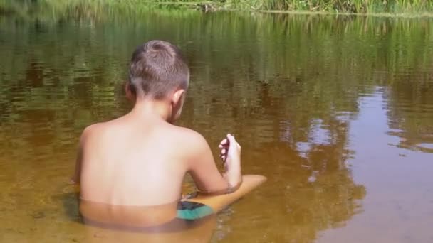 Un niño se sienta en una posición de loto en un río poco profundo al atardecer. 4K. De cerca. — Vídeo de stock