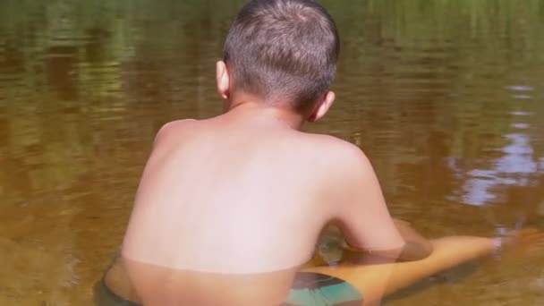 Achteraanzicht van een kind zittend op zand tot aan de taille in een transparante, ondiepe rivier — Stockvideo