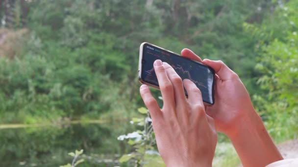 Un bărbat analizează graficul unei criptomonede într-un smartphone din natură. 4K — Videoclip de stoc