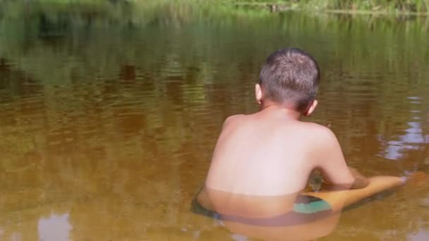 Vista posteriore di un bambino seduto sulla sabbia fino alla vita in un fiume trasparente e poco profondo — Video Stock