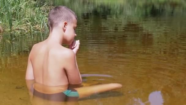 Een kind zit diep in de taille in een ondiepe rivier, bijtende nagels bij zonsondergang. 4K. Sluiten. — Stockvideo