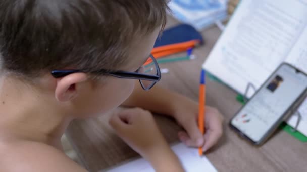 Školák v brýlích píše s perem, hledá v chytrém telefonu. 4K. Zpomalený pohyb — Stock video