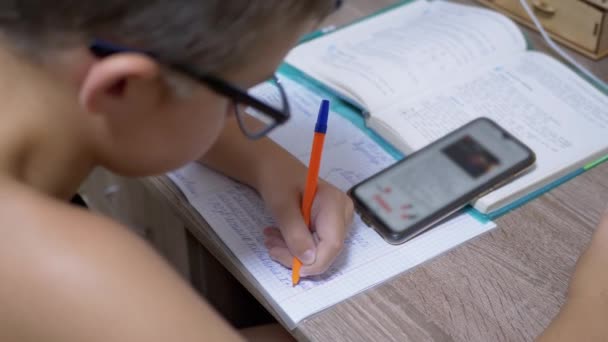 A szemüveges iskolás fiú tollal ír, a Smart Phone-t nézi. 4K. Lassú mozgás. — Stock videók
