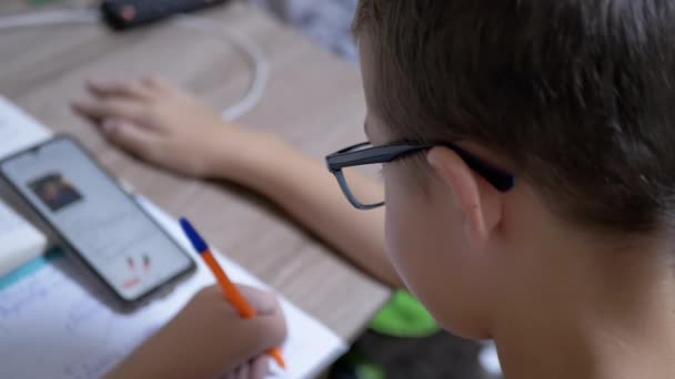 Schoolboy dans les lunettes de vue écrit avec stylo, à la recherche dans le téléphone intelligent. 4K. Mouvement lent — Video