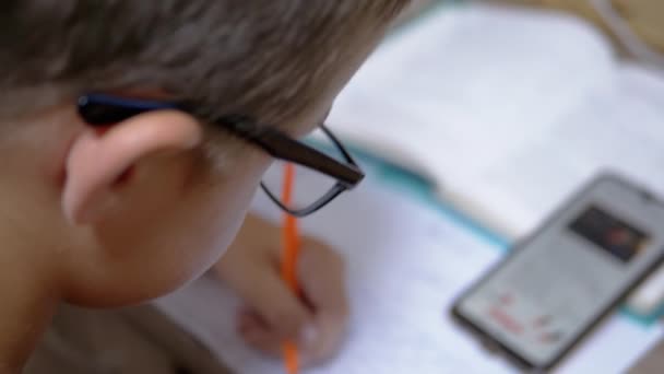 Schooljongen in brillen Schrijft met pen, kijkend in Smart Phone. 4K. Langzame beweging — Stockvideo