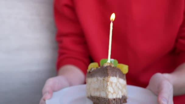 Μια γυναίκα κρατά ένα πιάτο με ένα κομμάτι τούρτα γενεθλίων και ένα φωτισμένο κερί. Εστίαση — Αρχείο Βίντεο