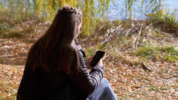 Fiatal nő szöveges üzenetet olvas egy okostelefonon, ül a lehullott száraz levelek — Stock videók