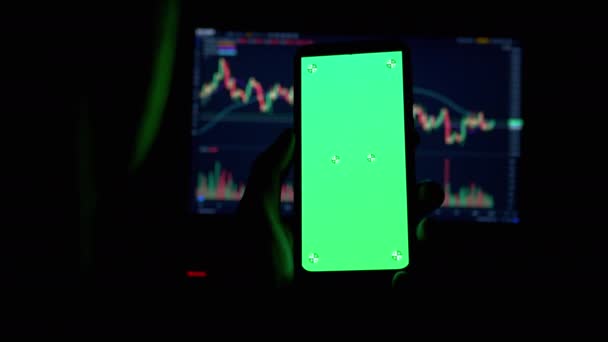 Трейдер держит смартфон с зеленым экраном в руках в темной комнате. 4K — стоковое видео