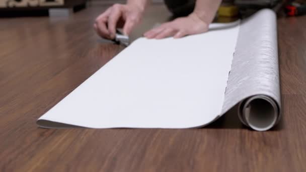Masculino, sentado no chão, corta uma tira de papel de parede para colar com uma faca — Vídeo de Stock