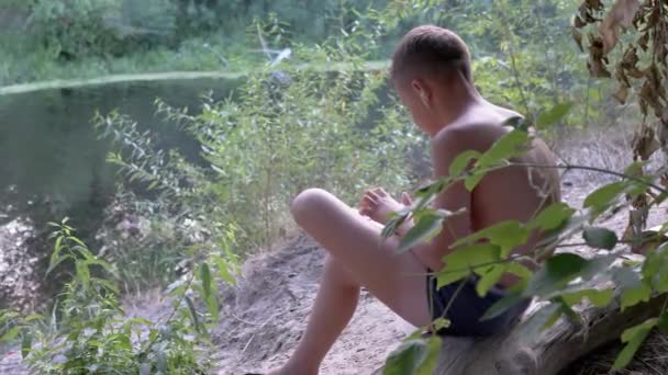 Kind spielt mit Kopfhörern auf dem Smartphone, sitzt auf einem trockenen Baumstamm im Wald. 4K — Stockvideo