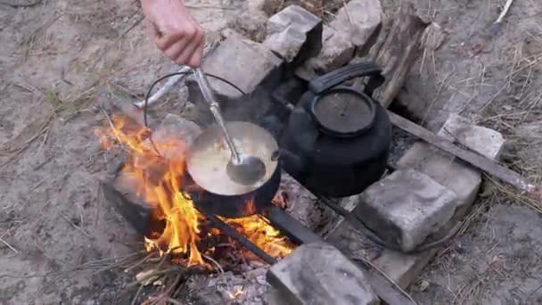 Homme Préparer la nourriture dans une casserole sur un feu de joie dans les bois à l'extérieur. 4K. Gros plan — Video