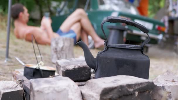 Old Black Kettle is Warming up by a Fire Against background of a Resting Tourist — Stock Video