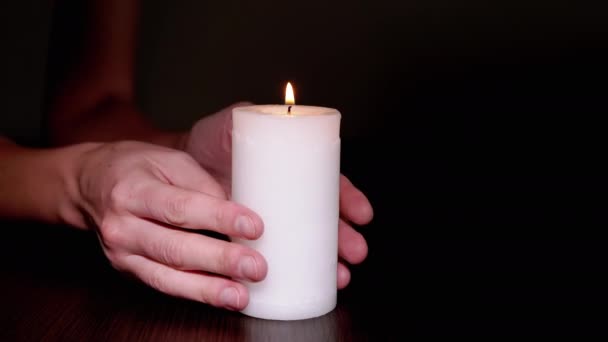 Feminino segura uma vela de cera branca queimando em uma mesa em um quarto escuro com as mãos — Vídeo de Stock