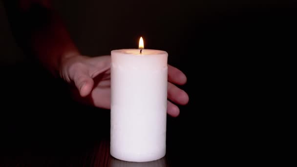 Een vrouwelijke hand neemt een brandende witte kaars van een tafel in een donkere kamer. Sluiten. — Stockvideo