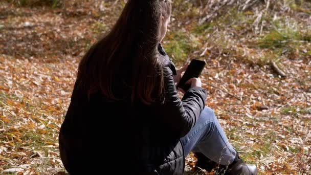 Fiatal nő szöveges üzenetet olvas egy okostelefonon, ül a lehullott száraz levelek — Stock videók