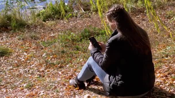 Jovem mulher lendo mensagem de texto em um smartphone, sentado em folhas secas caídas — Vídeo de Stock