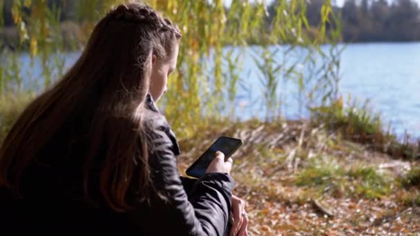 Fiatal nő szöveges üzenetet olvas egy okostelefonon, ül a lehullott száraz levelek — Stock videók