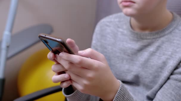 Niño se sienta en una silla, sostiene un teléfono inteligente en las manos, juega en el dormitorio. De cerca. — Vídeo de stock