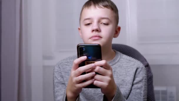 Niño sonriente se sienta en una silla, sostiene un teléfono inteligente en las manos, lee SMS en la pantalla — Vídeo de stock