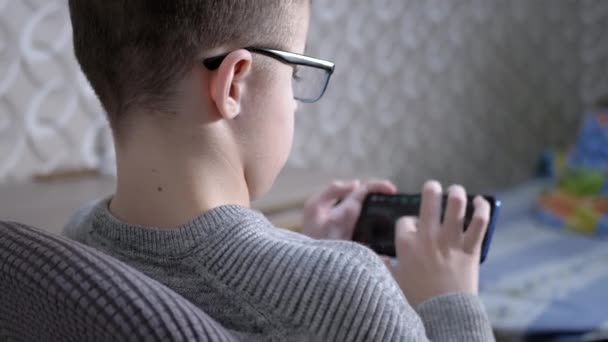 Уставший ребенок в очках сидит в кресле, играет в видеоигры на смартфоне. 4K — стоковое видео