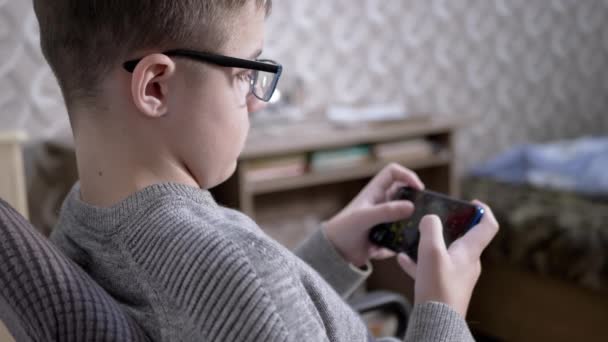 Zmęczone dziecko w okularach siedzi na krześle, gra w gry wideo na smartfonie. 4K — Wideo stockowe