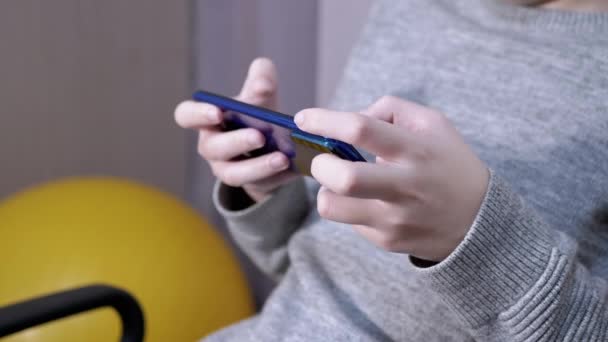 Criança senta-se em uma cadeira, segura um smartphone em mãos, joga no quarto. Fechar — Vídeo de Stock