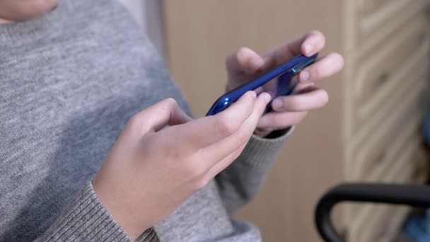 L'enfant s'assoit dans une chaise, tient un smartphone dans les mains, joue dans la chambre à coucher. Gros plan — Video