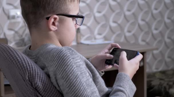 Criança cansada em óculos Senta-se em uma cadeira, joga videogames em um smartphone. 4K — Vídeo de Stock