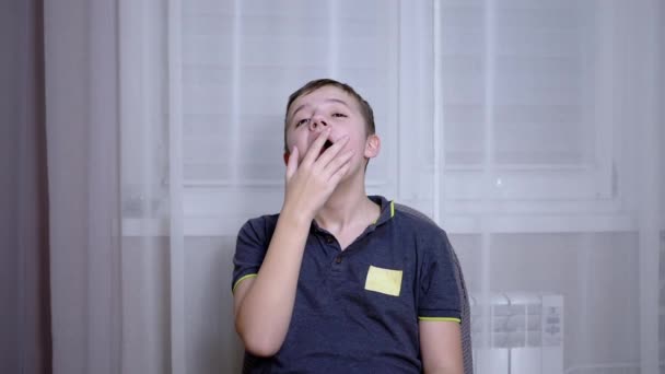 Álmos gyermek ásít, eltakarja a száját a kezével, ül egy székben a szobában — Stock videók