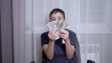 Mutlu Çocuk 'un elinde bir sürü 100 Dolarlık banknot, sandalyede oturuyor. Kapat.