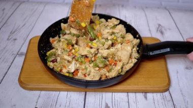 Kızarmış tavuk eti ile sebzeleri kızartma tavasında karıştıran kadın el. Renkli sebzelerle karıştırılmış parlak buğulanmış fileto parçaları. Bezelye, fasulye, mısır, havuç. Kesme tahtası. Tahta spatula.