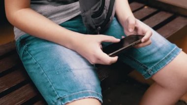 Park Outdoor 'da akıllı telefonda video oyunları oynayan, Bench' te oturan bir çocuk. Dinlenen çocuk elinde bir tablet, elinde bir cep telefonu tutuyor ve parmaklarıyla ekrana dokunuyor. Sosyal medya bağımlılığı.