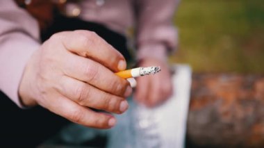 Woman Holding a Cigarette in Hands Sitting in Nature on Sun. Hands close-up. Smoke a fuming cigarette butt outdoors in the woods. Bad habit. Addiction. Nicotine, ash, poison. Unhealthy lifestyle.