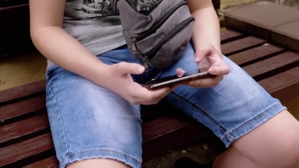 子供が公園の屋外でスマートフォンでビデオゲームをプレイ ベンチに座って 安静時の少年はタブレットを持ち 携帯電話を手に取り 指で画面に触れる ソーシャルメディア中毒 — ストック動画