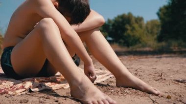 Sıkıcı Çıplak Çocuk Örtüye Oturur, Kumsalda Dal Kum Çizer. Çıplak bacaklar, kollar, can sıkıntısı üzerine çizim yapan yalnız ve yorgun bir çocuk. Sıcaklık, açık hava eğlencesi, yaz. Doğa.