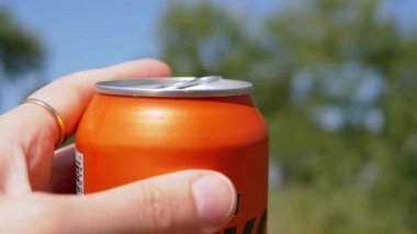 Doğanın bulanık zemininde Açık Teneke Bira Kutusu Tutan Kadın El. Renkli resimli doğa, açık hava, park, orman. Mavi gökyüzü, yeşil ağaçlar. Susadım. Alkol iç. - Şerefe. Dinleniyor. Hafta Sonu.