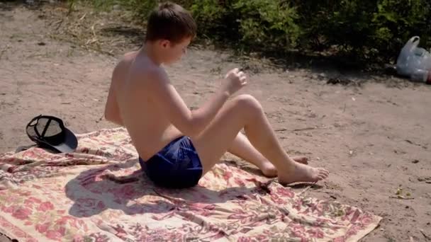 无聊的孩子坐在沙滩上的小木屋上 在炎热的阳光下在海滩上晒日光浴 疲倦的男孩低着头 在灼热的阳光下在大自然中歇息 — 图库视频影像