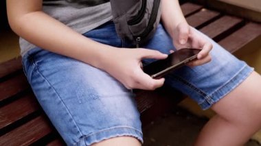 Park Outdoor 'da akıllı telefonda video oyunları oynayan, Bench' te oturan bir çocuk. Dinlenen çocuk elinde bir tablet, elinde bir cep telefonu tutuyor ve parmaklarıyla ekrana dokunuyor. Sosyal medya bağımlılığı.