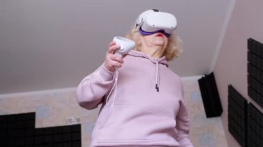 3D Gözlüklü Kadın Oyun Kontrolörleriyle Ses geçirmez Odada Oynuyor. Beyaz miğferli yaşlı oyuncu kadın, VR test cihazıyla el sallıyor. Sanal gerçeklik. Cihaz. Yapay zeka. 4K.