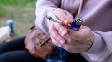 Elinde sigara tutan kadın ve doğada güneşin altında oturan bir çakmak. Dışarıda sigara içen bir kadın, park eder. Sigara içiyorum. Kötü alışkanlık. Bağımlılık. Sağlıksız yaşam tarzları. Stres yüzünden. Dinleniyor.